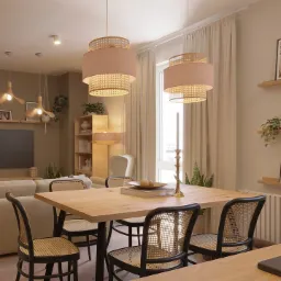 Jasne wnętrze jadalni z drewnianym stołem, krzesłami z rattanowym oparciem, dwoma wiszącymi lampami z różowymi abażurami i rattanowymi elementami oraz dekoracjami roślinnymi na półkach.