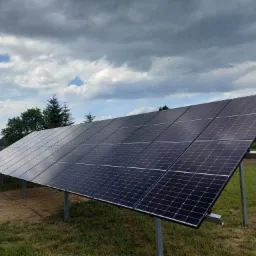 Rząd paneli słonecznych zamontowanych na metalowych słupkach na trawiastym polu, z pochmurnym niebem w tle.