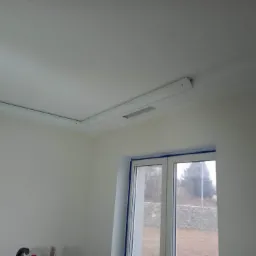 Wykończone wnętrze: gładka, biała ściana i sufit z zamontowaną listwą oświetleniową z wbudowanym nawiewnikiem, okno zabezpieczone taśmą malarską.