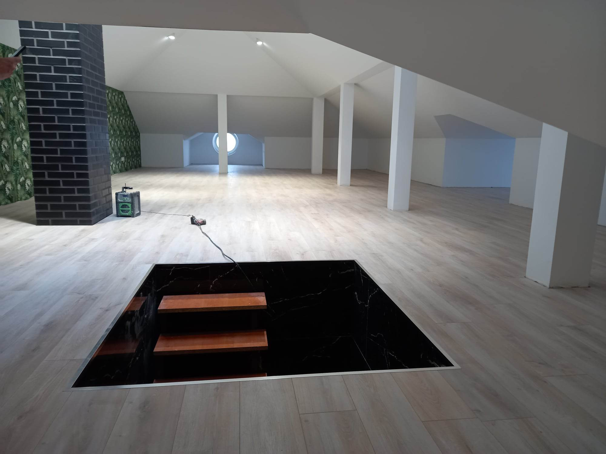 Jasne panele podłogowe w przestronnym poddaszu z czarnym, marmurkowym wykończeniem otworu ze schodami prowadzącymi w dół; widoczny fragment ściany z cegły i tapetą w roślinny wzór.