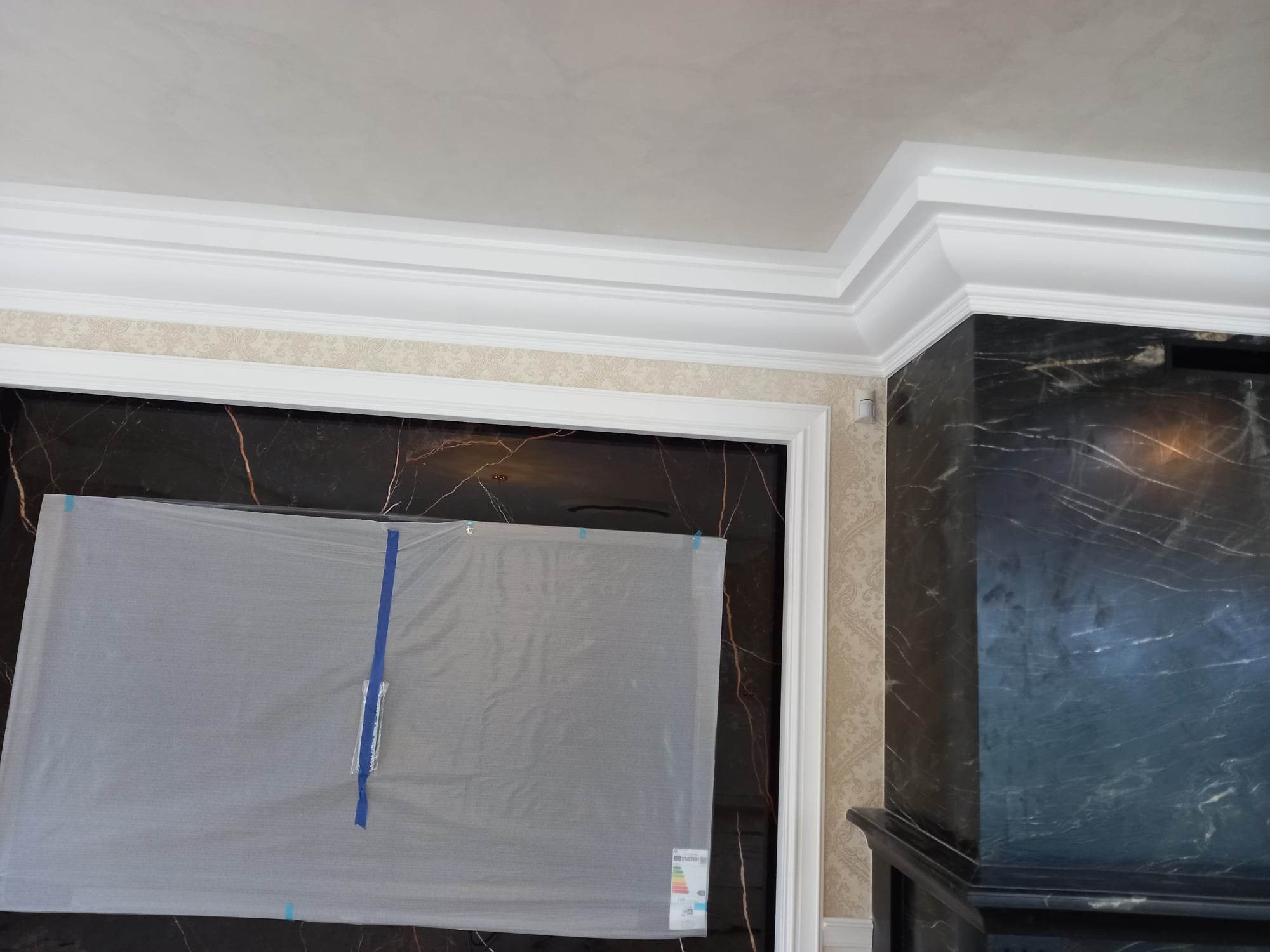 Fragment wnętrza z marmurową ścianą, tapetą w delikatny wzór i białymi listwami dekoracyjnymi pod sufitem, zabezpieczona folią ściana.