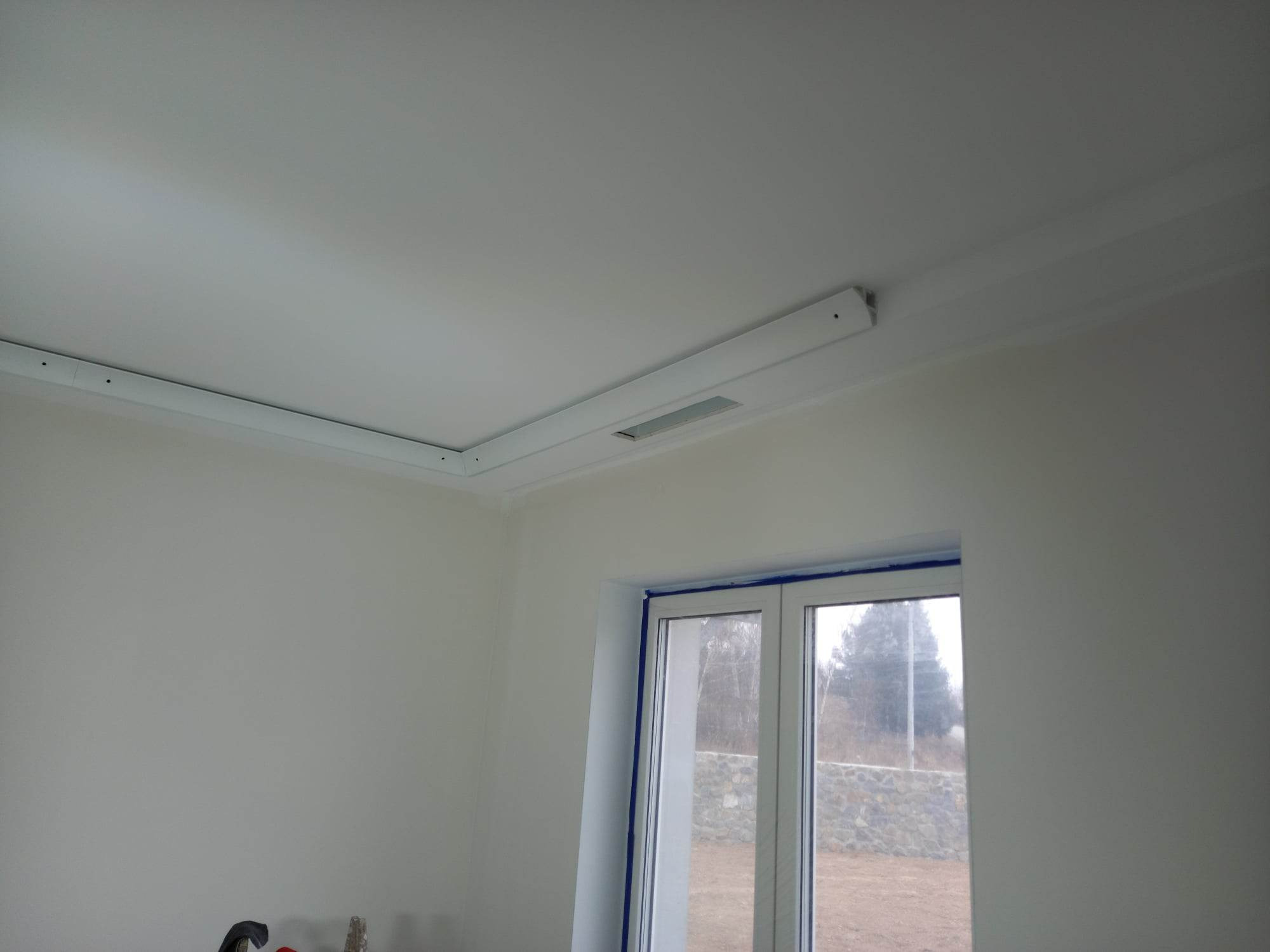 Wykończone wnętrze: gładka, biała ściana i sufit z zamontowaną listwą oświetleniową z wbudowanym nawiewnikiem, okno zabezpieczone taśmą malarską.