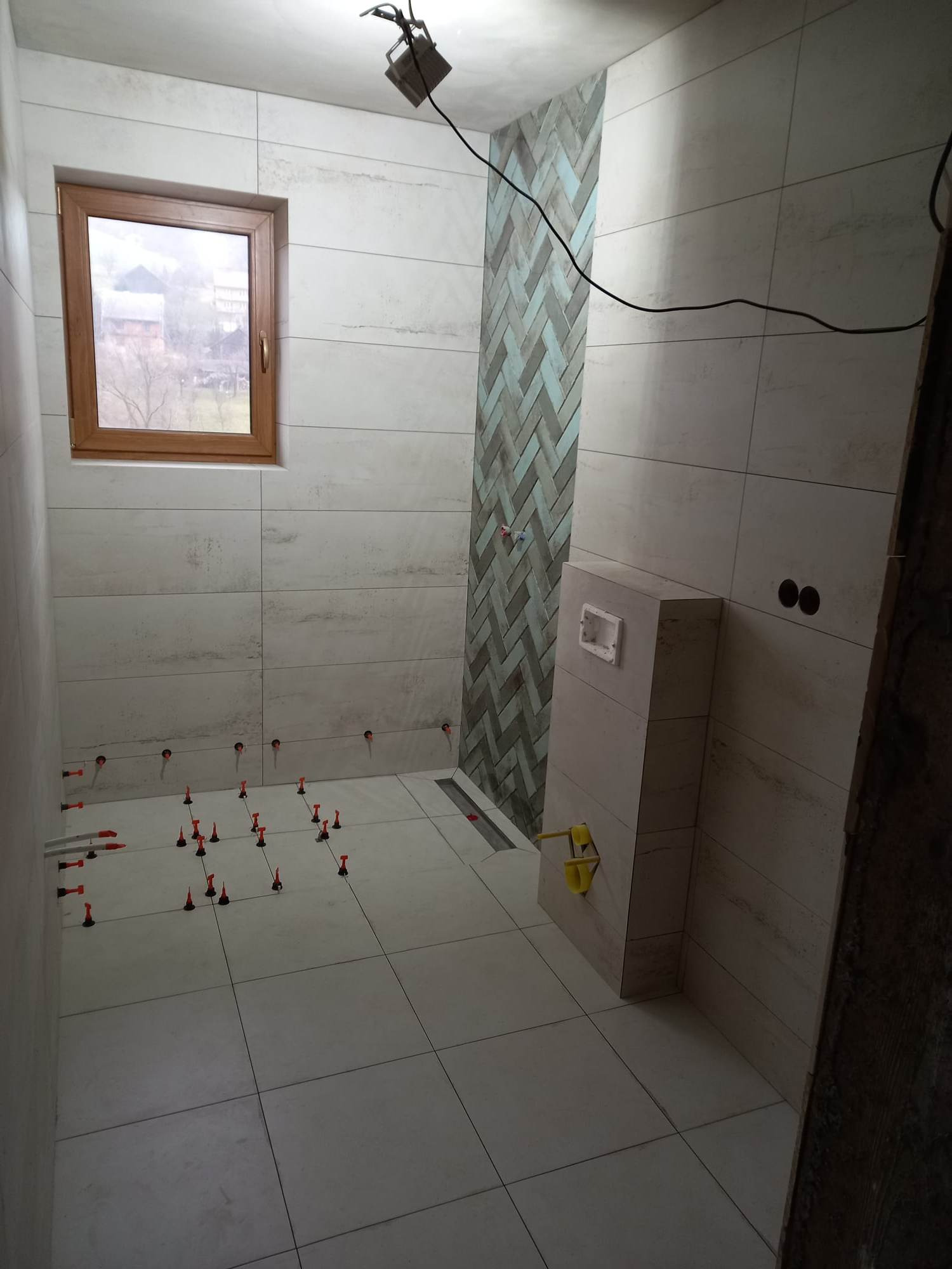 Łazienka w trakcie wykańczania: jasne, poziome płytki imitujące drewno na ścianach, na podłodze kwadratowe płytki, rząd niebiesko-szarych płytek ułożonych w jodełkę, widoczne systemy poziomowania...