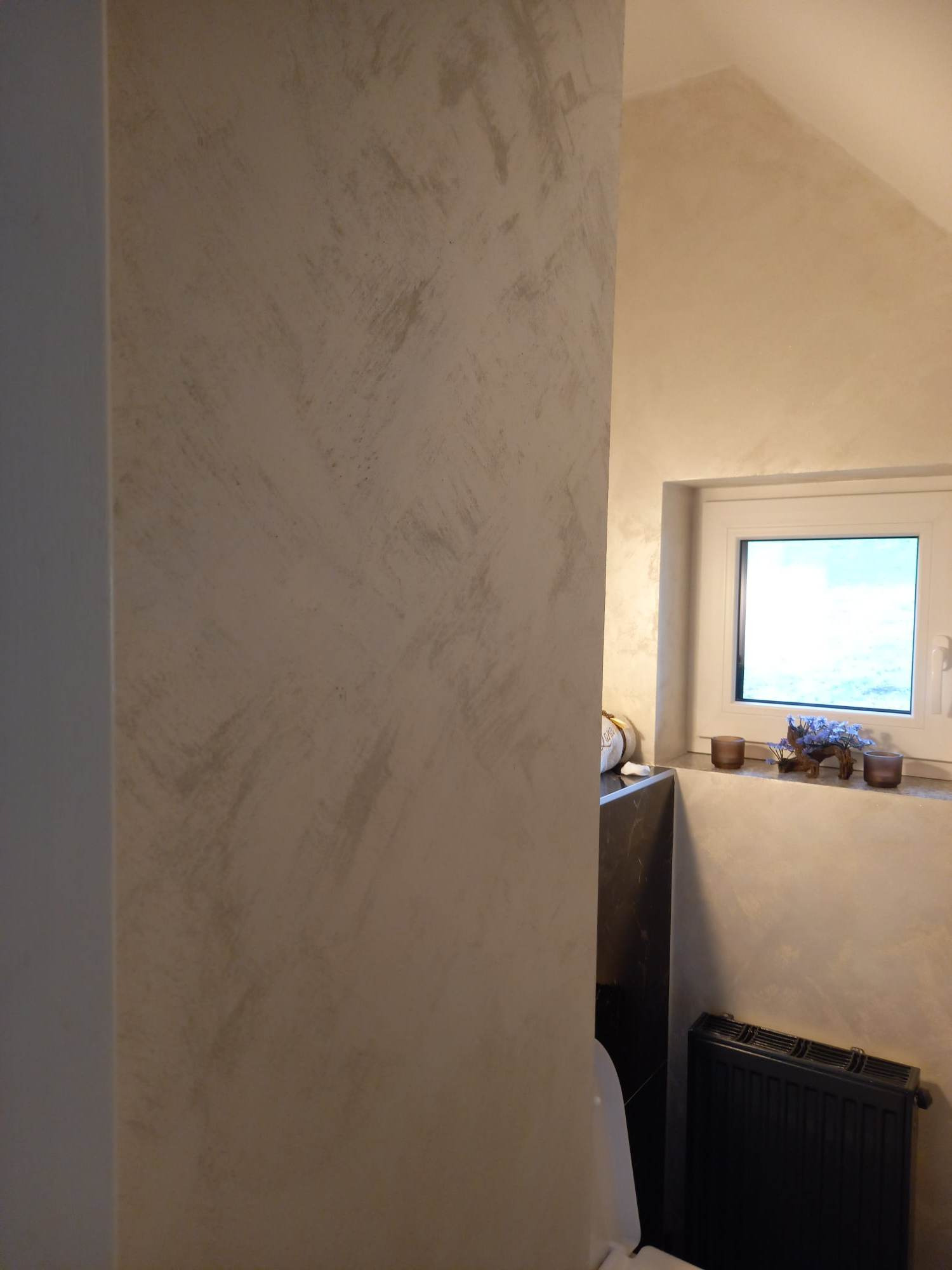 Ściana w łazience wykończona dekoracyjną, beżowo-szarą techniką malarską, widoczny fragment toalety, okno z dekoracjami i grzejnik.