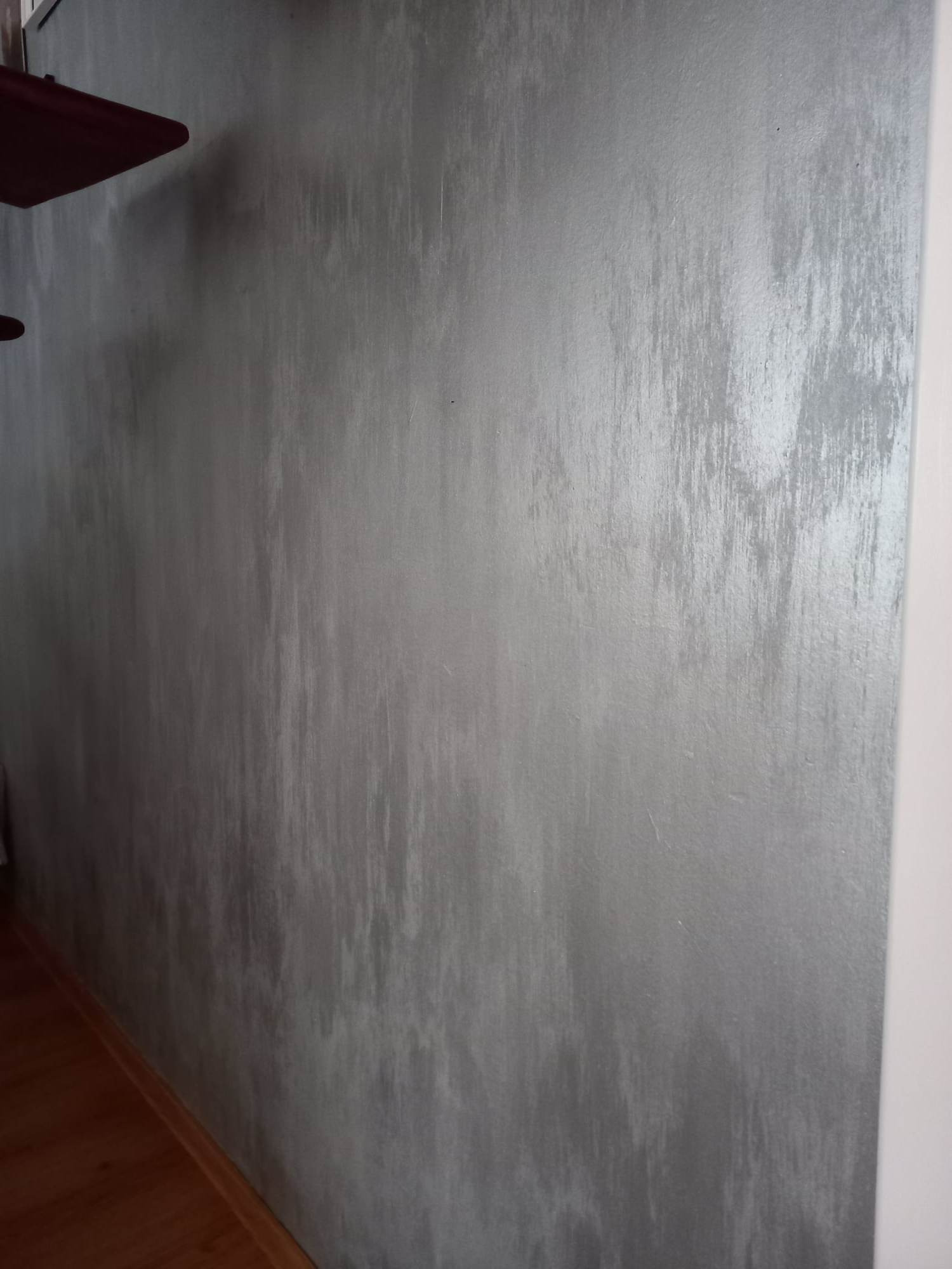 Ściana z dekoracyjną, srebrzystą powłoką, imitującą efekt przetarć i smug, widoczna z bliska, fragment brązowej półki w lewym górnym rogu, drewniana podłoga w dolnej części kadru.
