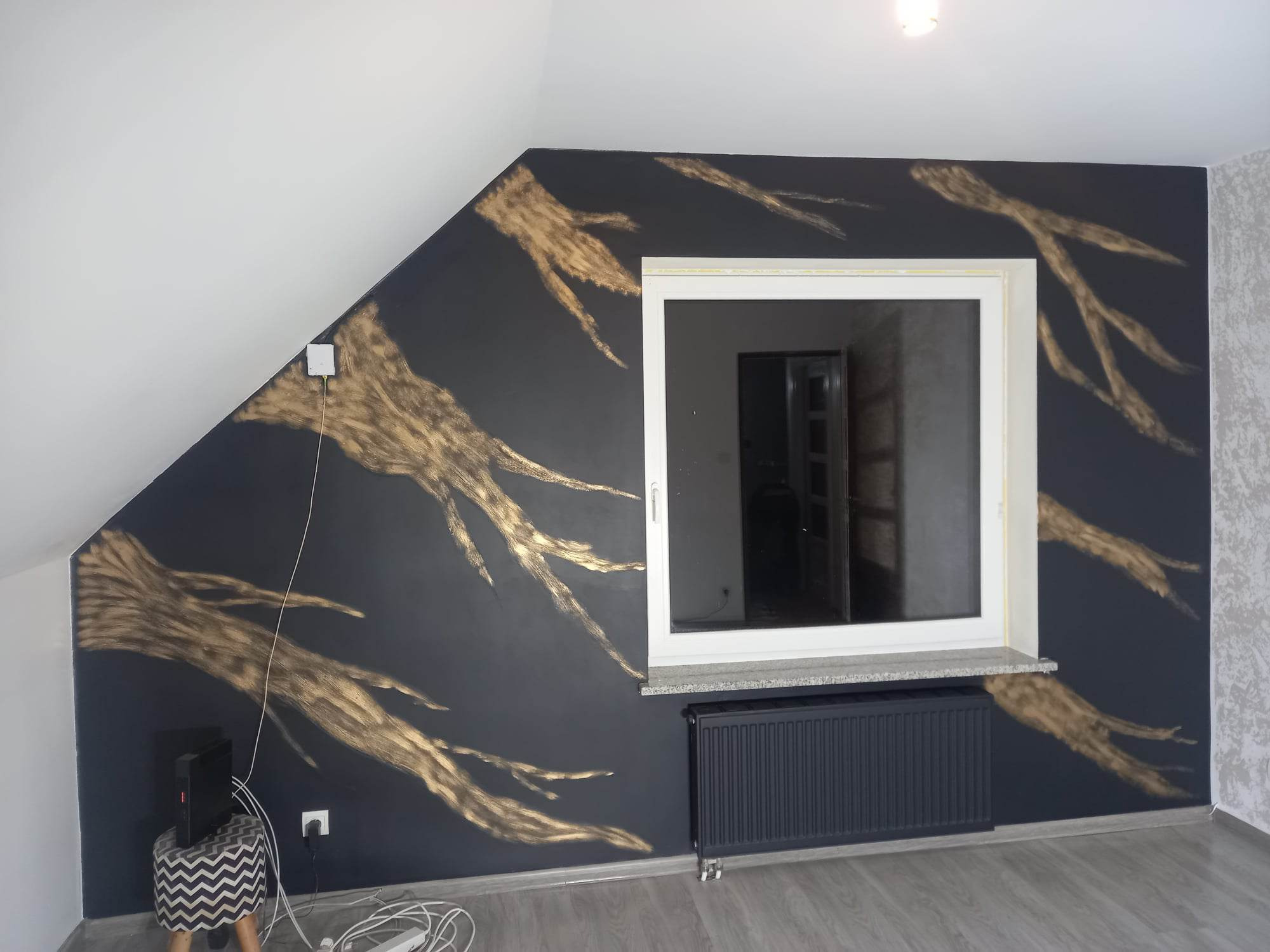 Ściana w pokoju na poddaszu pomalowana na ciemny kolor z ręcznie malowanymi złotymi wzorami przypominającymi gałęzie drzew, białe okno z szarym parapetem, grzejnik pod oknem, szara podłoga z paneli.