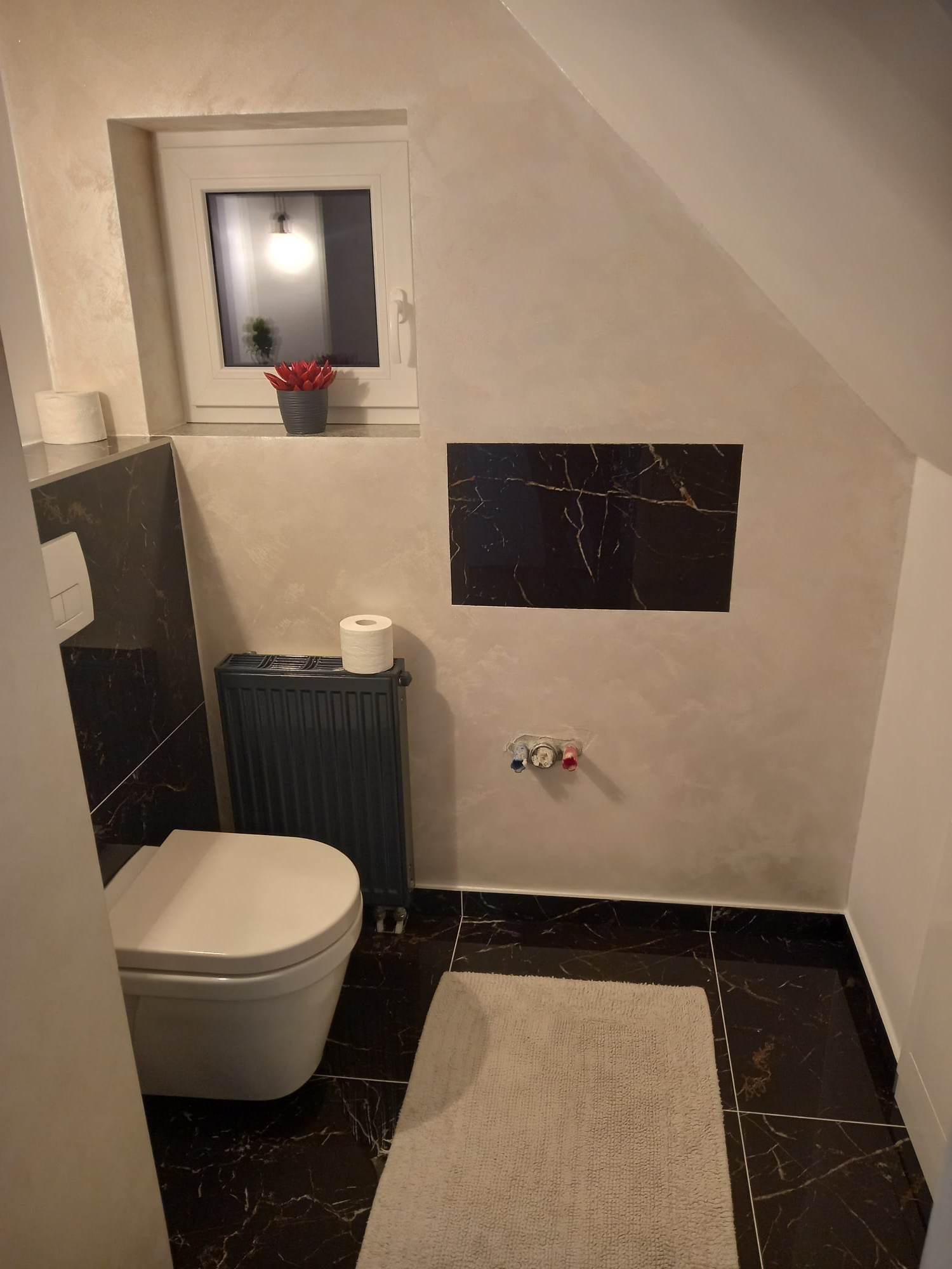 Wykończona łazienka na poddaszu w Nowym Sączu z czarnymi płytkami imitującymi marmur na podłodze i ścianie, gładzią na skośnej ścianie, grzejnikiem żeberkowym i wiszącą toaletą.