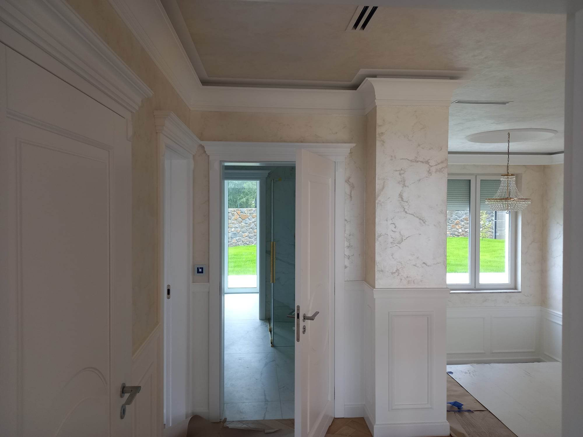 Eleganckie wnętrze po remoncie: marmurkowe ściany, biała stolarka drzwiowa i kolumna dekoracyjna z panelami, widoczny fragment parkietu i kryształowy żyrandol w tle.