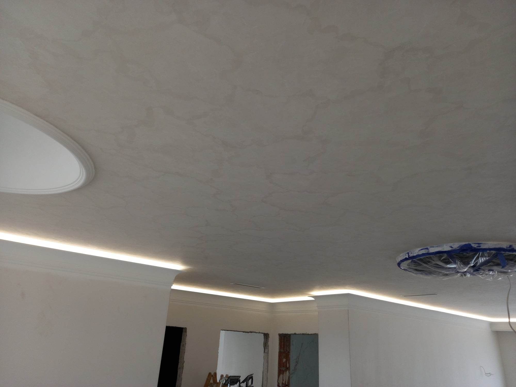 Wykończony sufit z dekoracyjną gładzią w odcieniach beżu, oświetlenie LED wzdłuż krawędzi, ozdobne listwy przysufitowe oraz zabezpieczony folią punkt oświetleniowy.