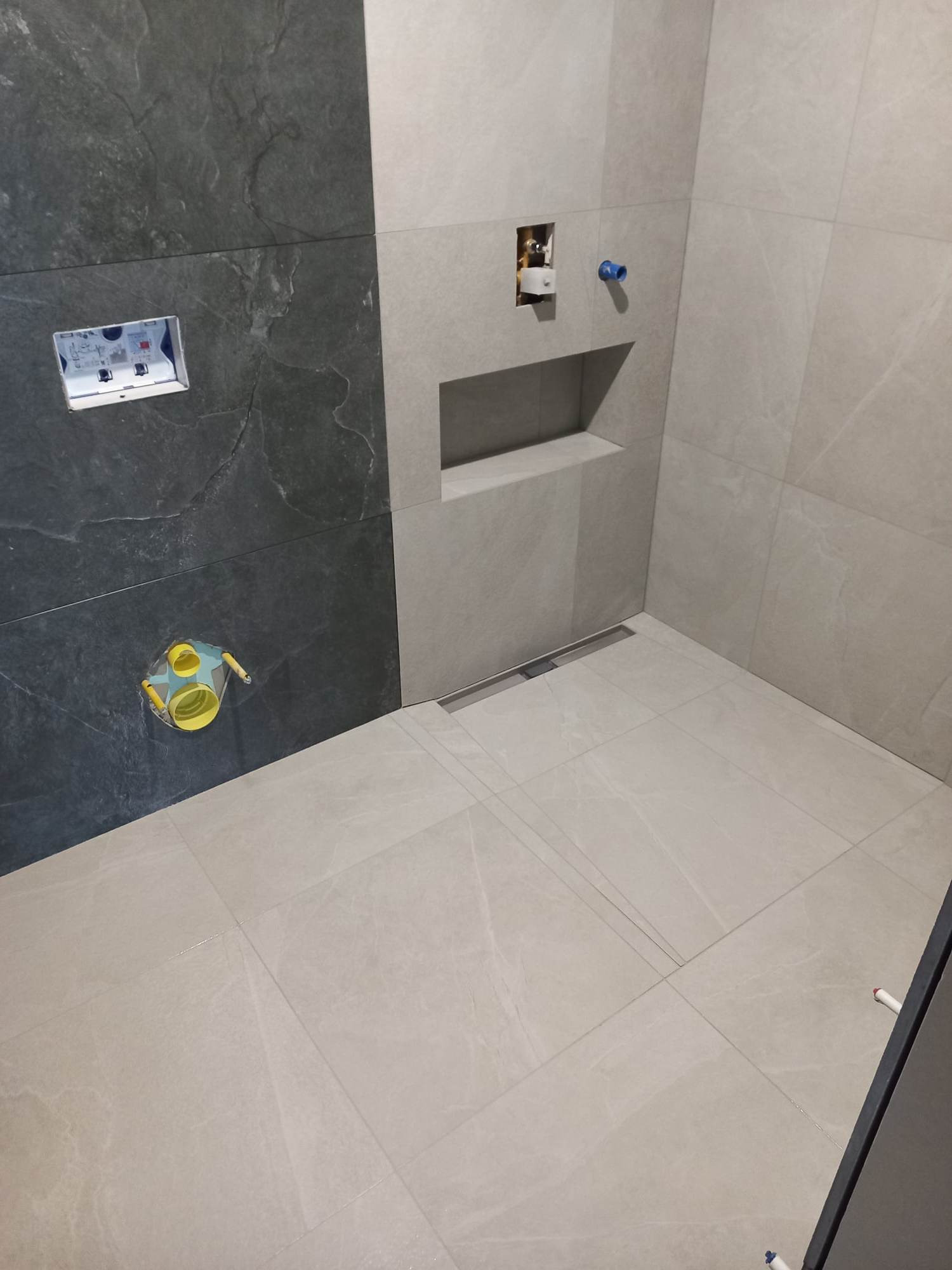 Wykończona łazienka z szarymi płytkami w dwóch odcieniach, widoczne podłączenia hydrauliczne i odpływ liniowy w podłodze.