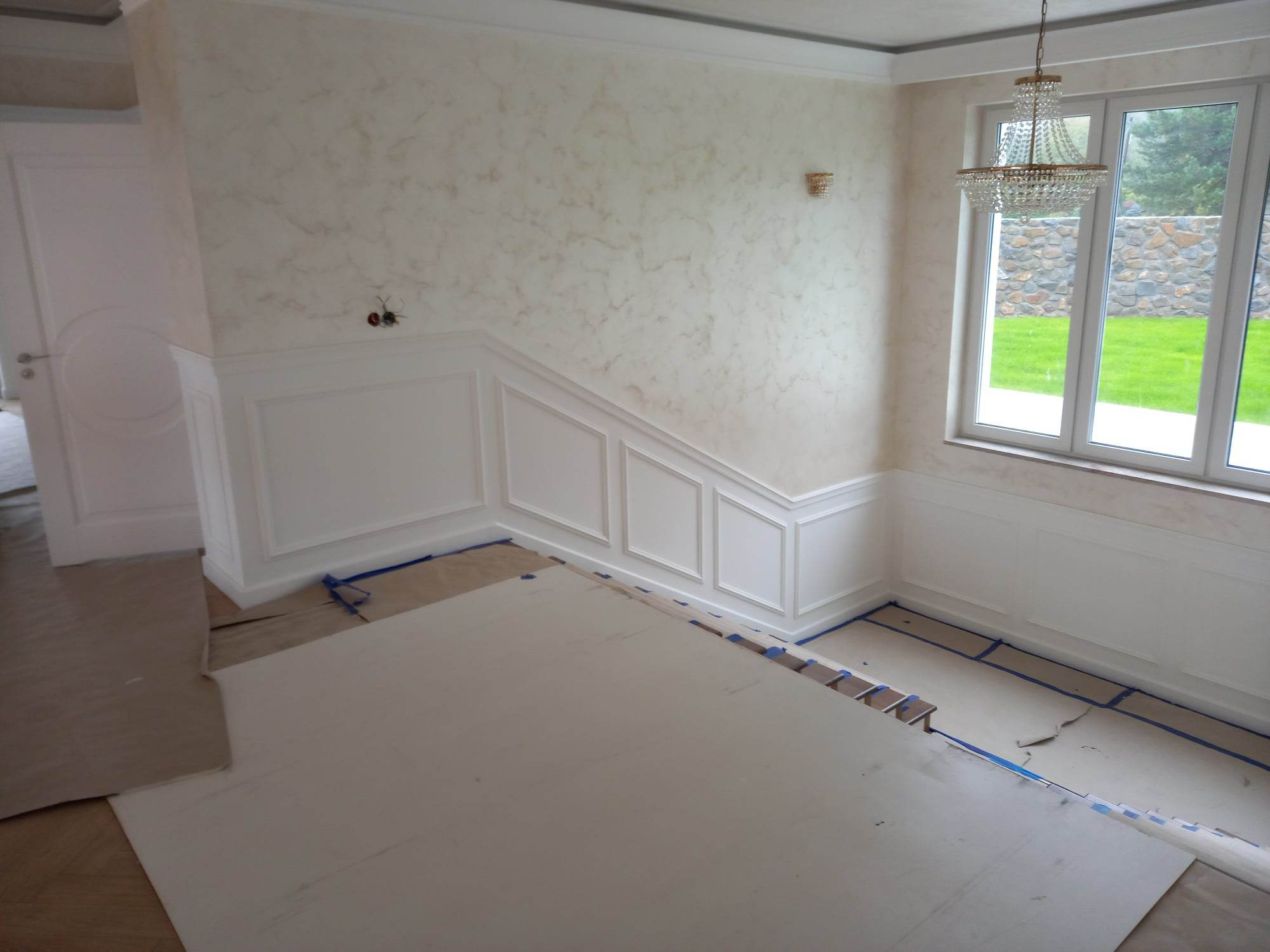 Pokój w trakcie prac wykończeniowych, ściany z dekoracyjną fakturą imitującą marmur, biała sztukateria na ścianach, podłoga zabezpieczona kartonem i taśmą malarską przed zabrudzeniem, widoczny...