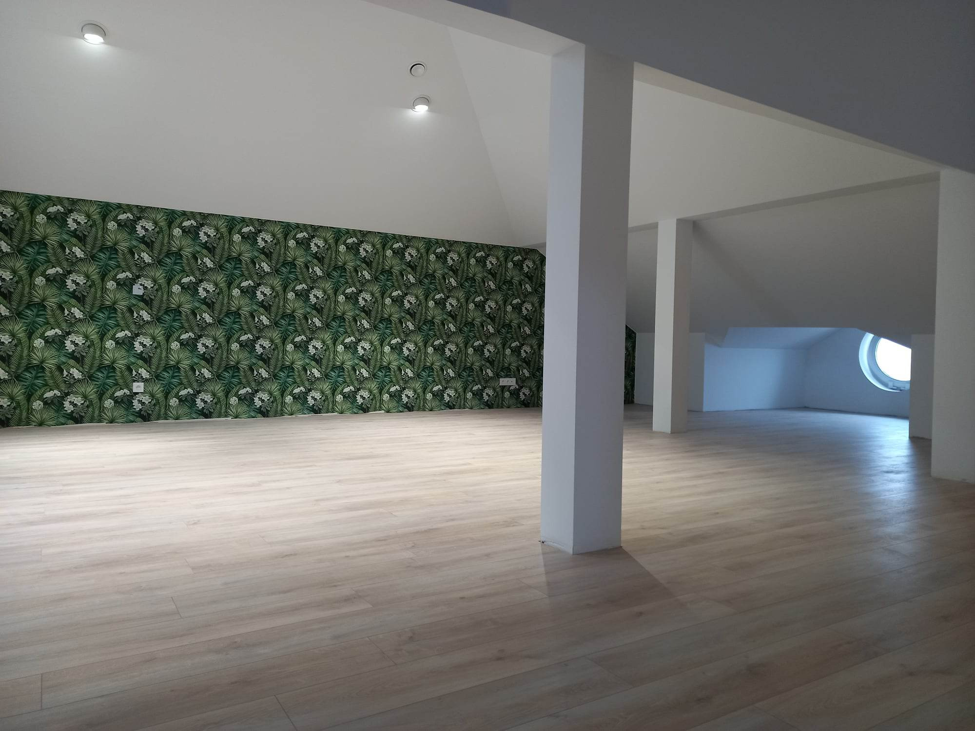 Jasne panele podłogowe w przestronnym pokoju z zieloną tapetą w tropikalne liście na jednej ścianie i białymi kolumnami podtrzymującymi sufit.
