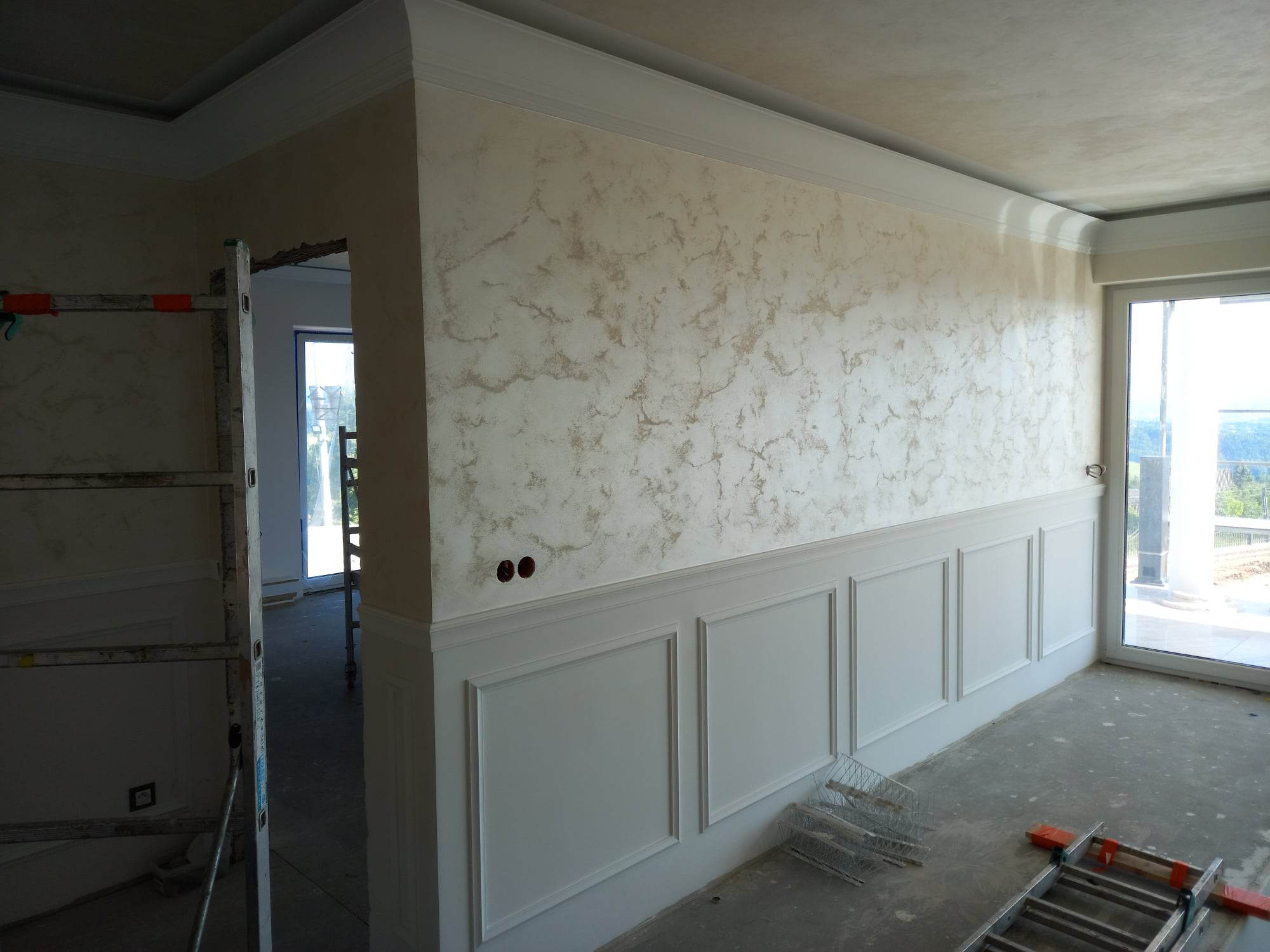 Wnętrze pokoju w trakcie prac wykończeniowych: ściany z dekoracyjną fakturą i białą boazerią, widoczne drabiny i narzędzia, surowy beton na podłodze.