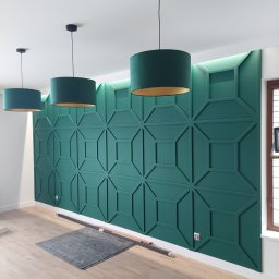 E&K BUD Krzysztof Smolarczyk - Zielona ściana dekoracyjna z geometrycznym wzorem, podświetlana od góry, uzupełniona trzema wiszącymi lampami z abażurami w odcieniach zieleni w pomieszczeniu w Kielcach.