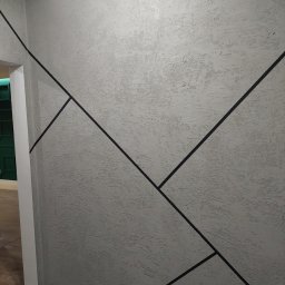 E&K BUD Krzysztof Smolarczyk - Szara ściana z dekoracyjną fakturą i geometrycznym wzorem z czarnych linii, zabezpieczona taśmą malarską przy podłodze i suficie.