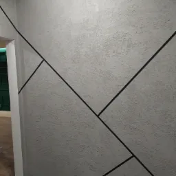 Szara ściana z dekoracyjną fakturą i geometrycznym wzorem z czarnych linii, zabezpieczona taśmą malarską przy podłodze i suficie.