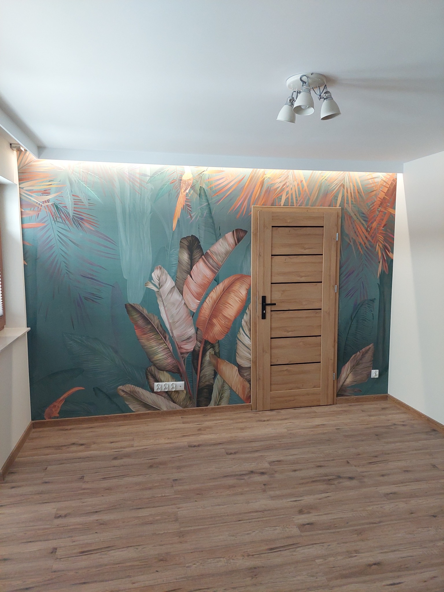 Wnętrze pokoju z drewnianą podłogą, ścianą z fototapetą w motywy tropikalne i zamontowanymi drewnianymi drzwiami z czarną klamką.