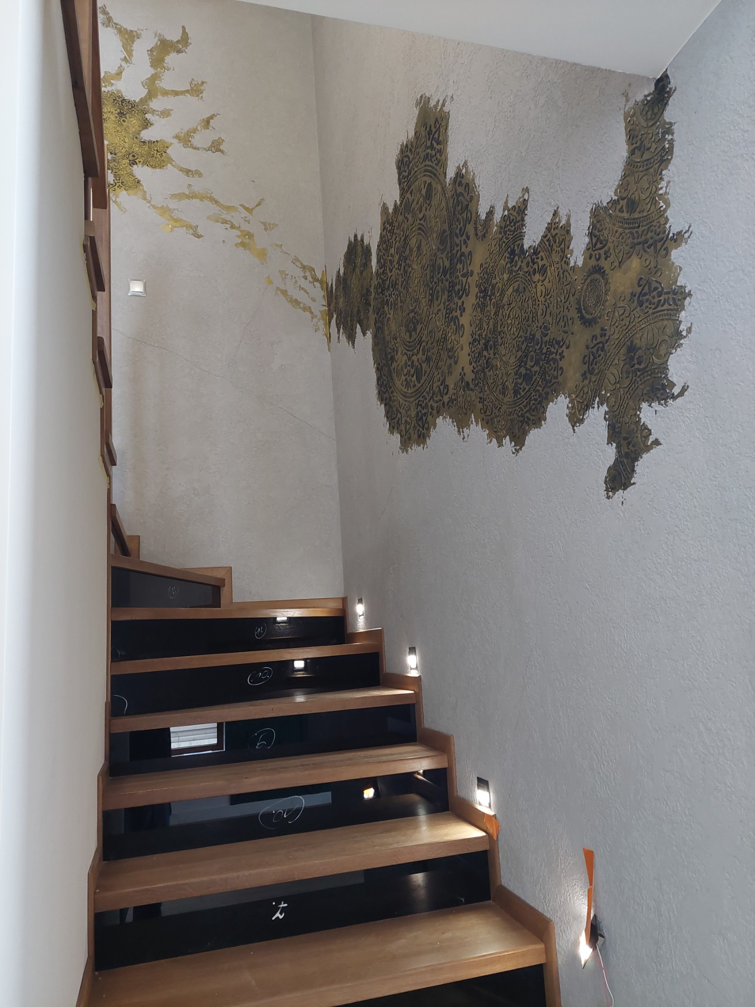 Klatka schodowa z drewnianymi schodami i oryginalną dekoracją ściany w kolorze złota i czerni.