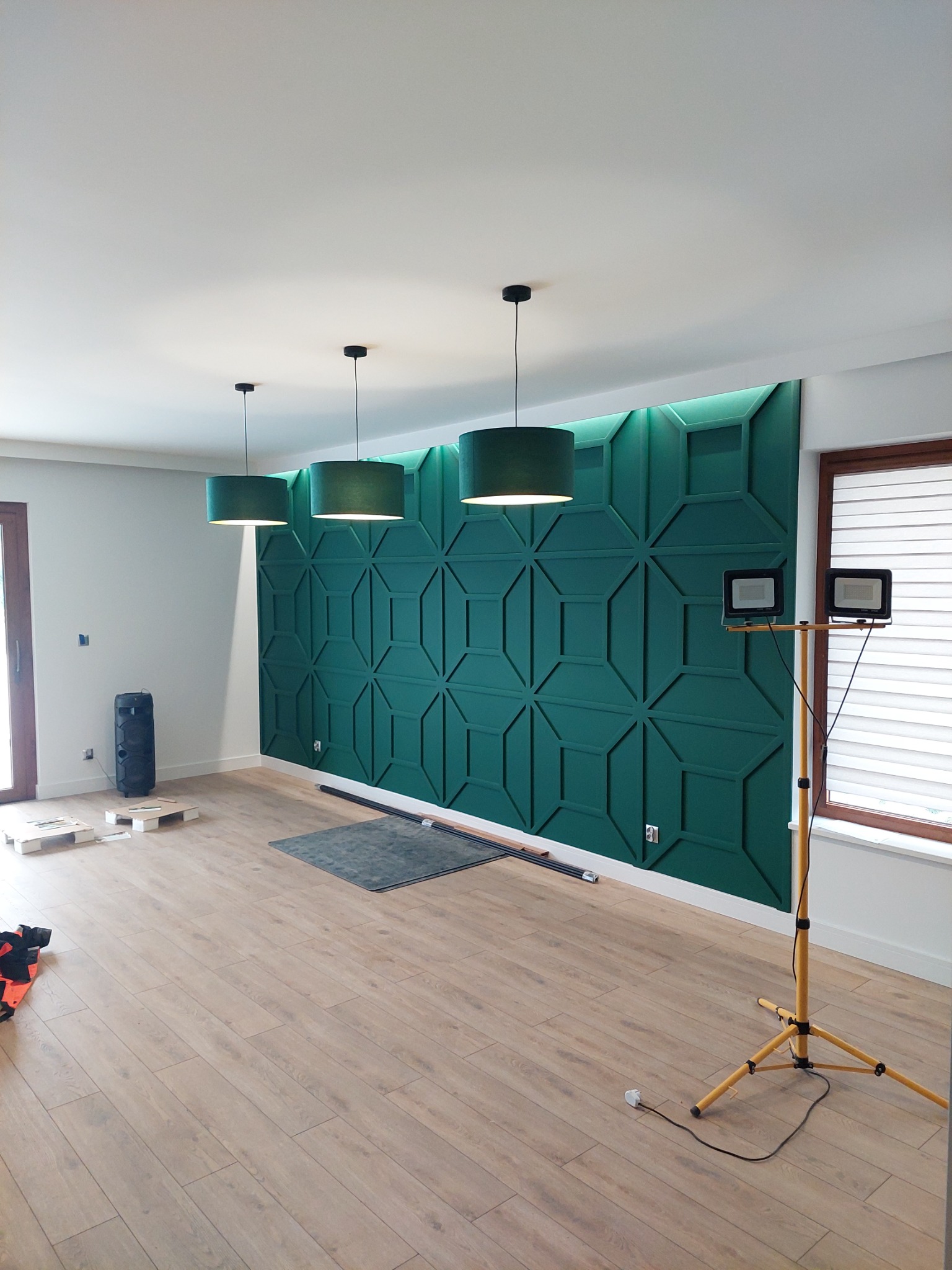 Pokój z zieloną, geometryczną ścianą dekoracyjną, oświetlony trzema wiszącymi lampami i dodatkowym reflektorem na statywie.