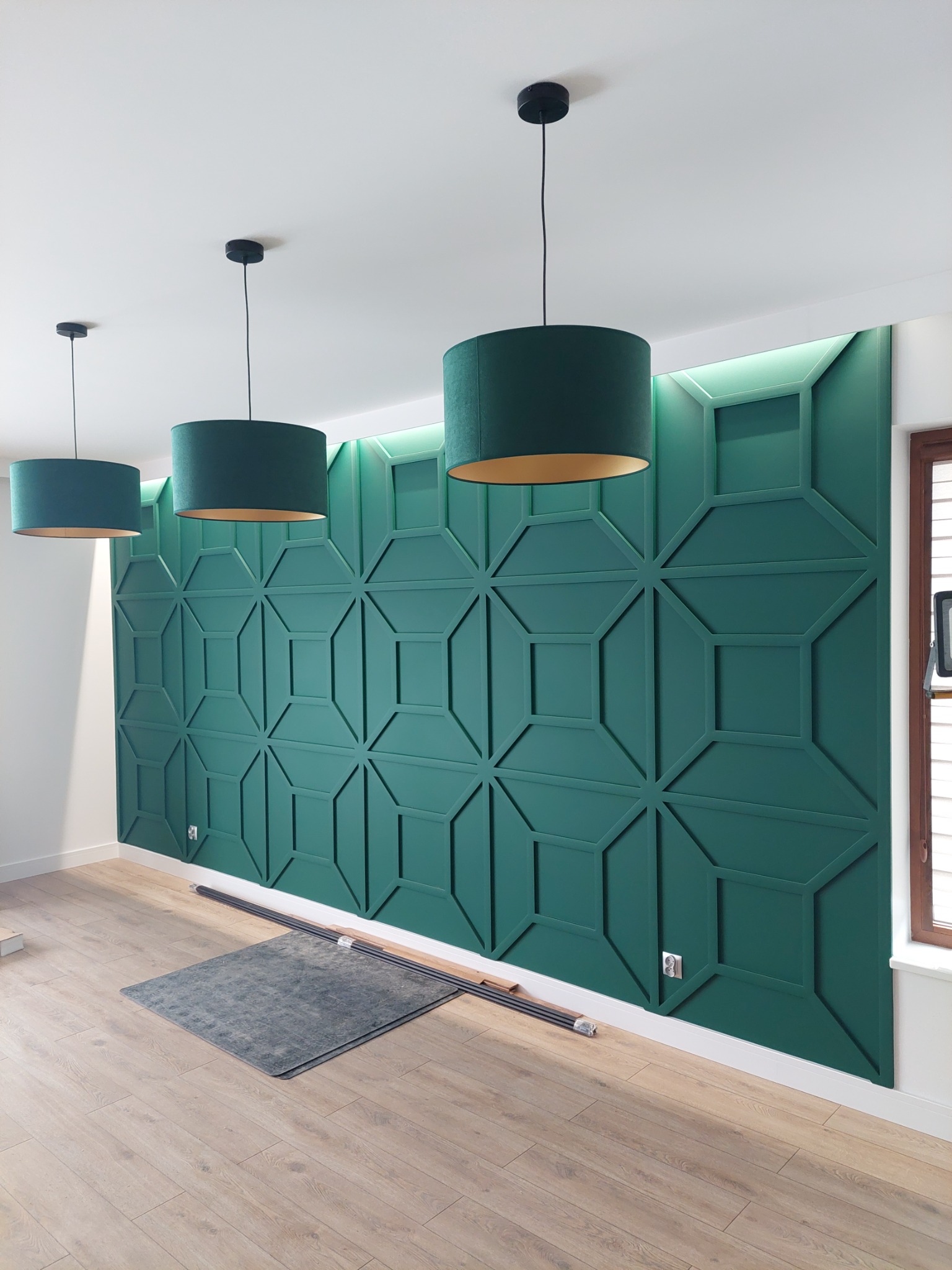 Zielona ściana dekoracyjna z geometrycznym wzorem, podświetlana od góry, uzupełniona trzema wiszącymi lampami z abażurami w odcieniach zieleni w pomieszczeniu w Kielcach.