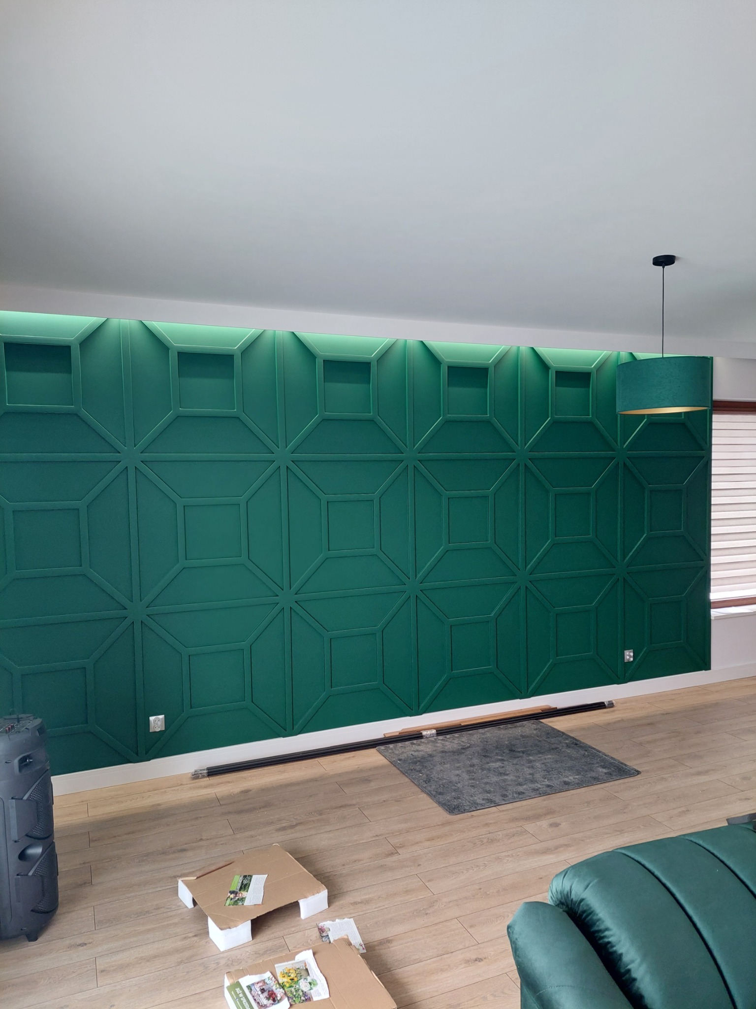 Szmaragdowa ściana dekoracyjna z podświetleniem LED na górze, wzór geometryczny kwadratów i rombów, fragment pokoju z lampą i podłogą drewnopodobną.