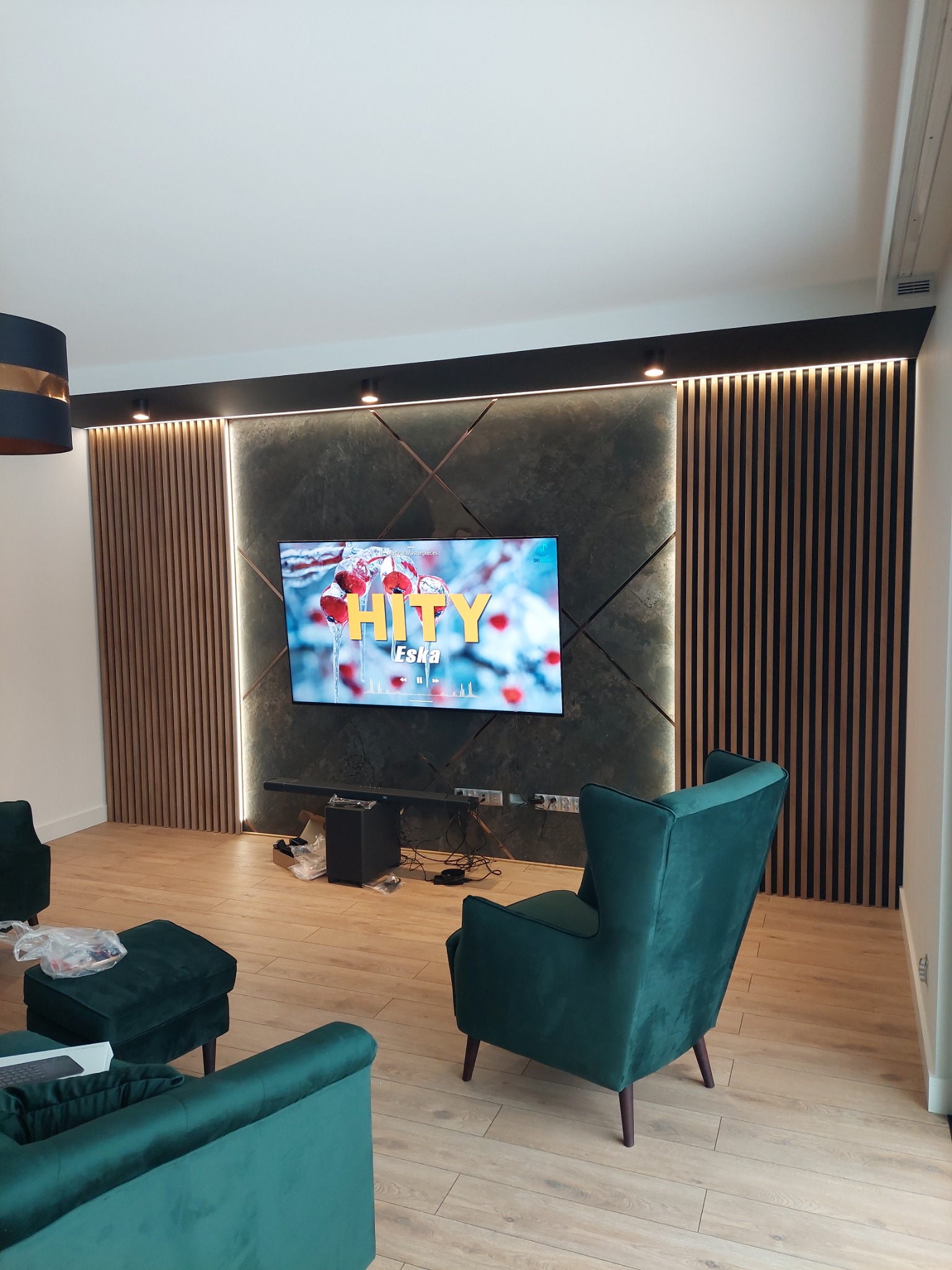 Nowoczesny salon z panelem ściennym z lameli drewnianych i zielonymi fotelami, oświetlenie punktowe nad telewizorem