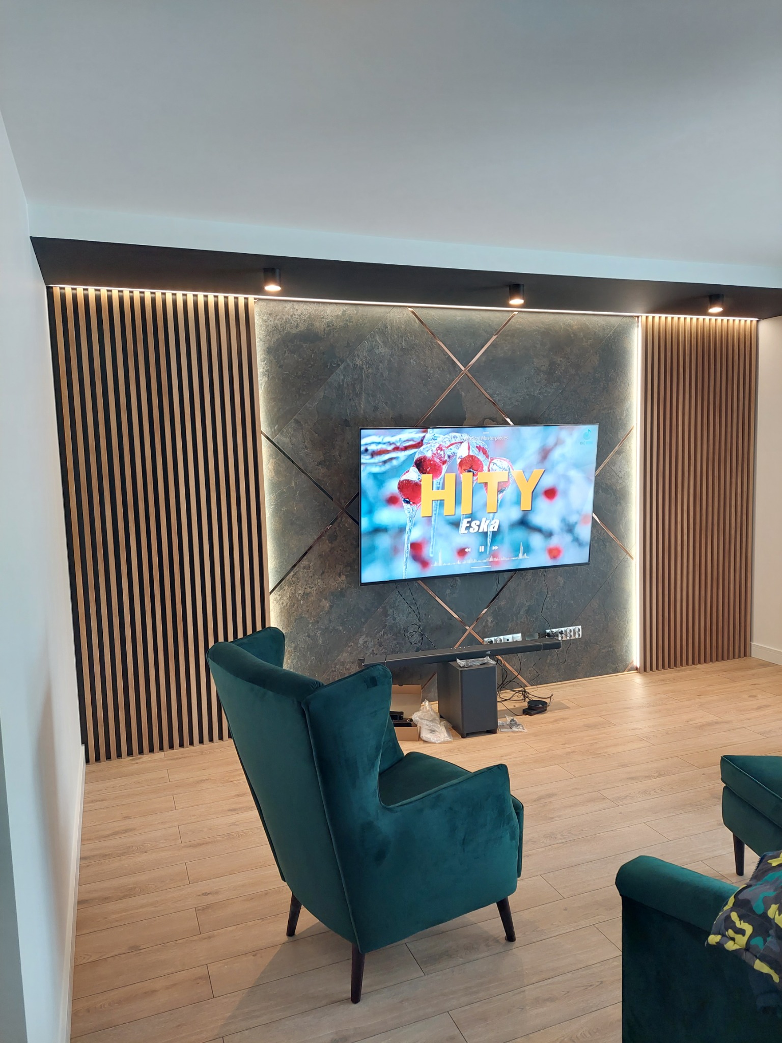 Nowoczesny salon z telewizorem na tle ściany z szarego kamienia, drewnianych paneli i miedzianych akcentów, oświetlenie punktowe, widoczne zielone fotele.