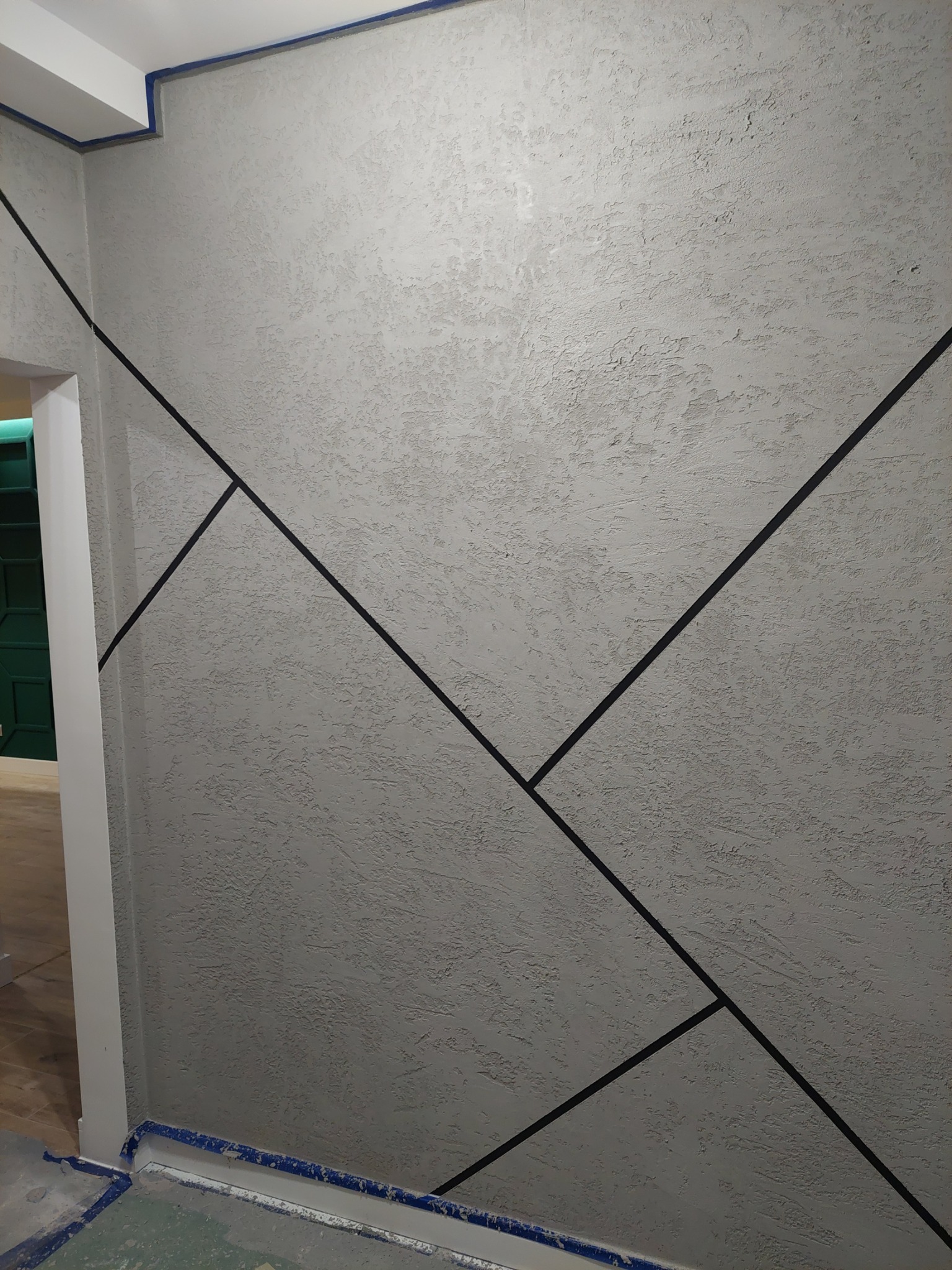 Szara ściana z dekoracyjną fakturą i geometrycznym wzorem z czarnych linii, zabezpieczona taśmą malarską przy podłodze i suficie.