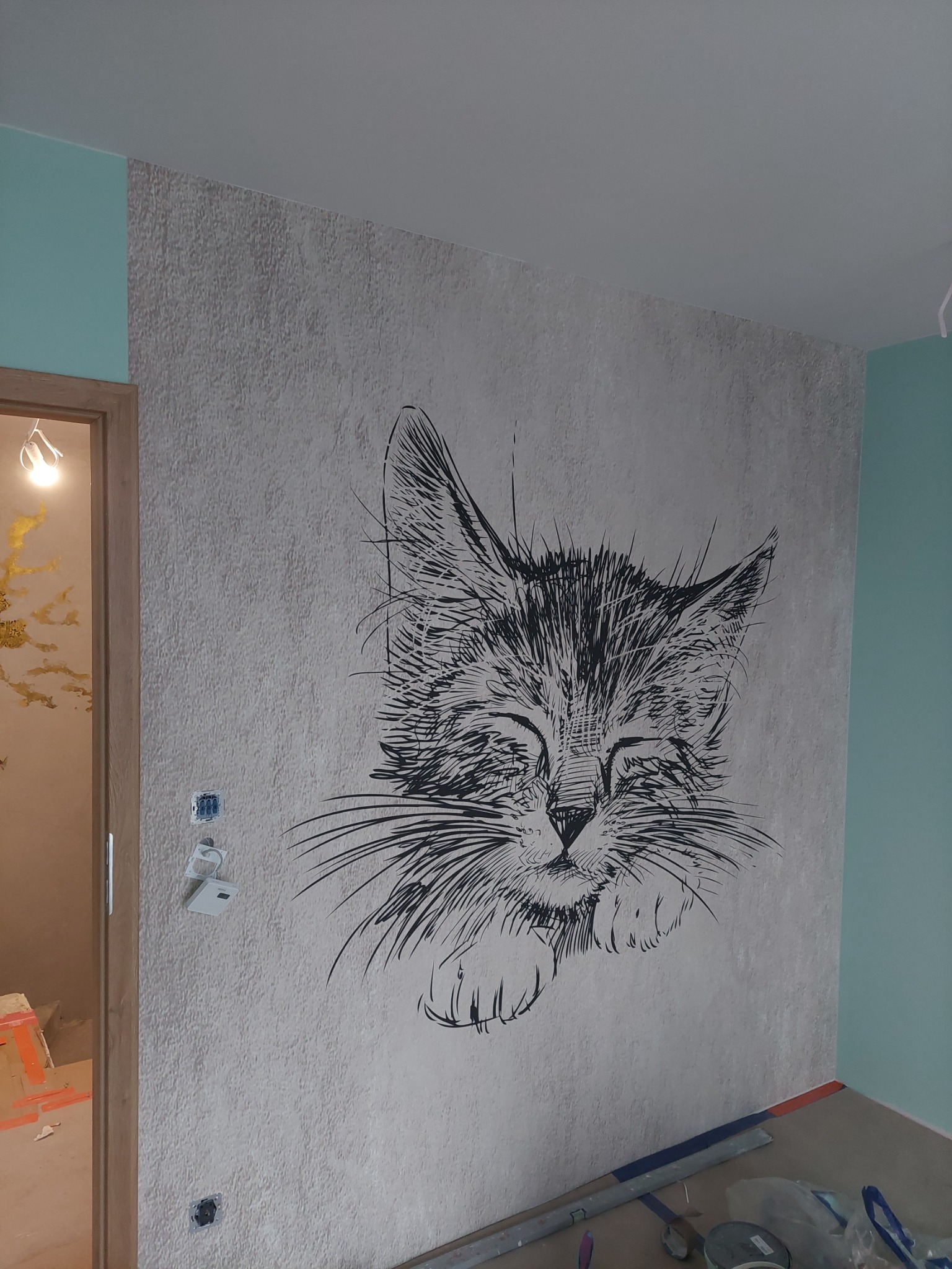 Ściana z szarą tapetą o fakturze z czarnym rysunkiem kota, widoczne przygotowania do wykończenia, zabezpieczenia na podłodze.
