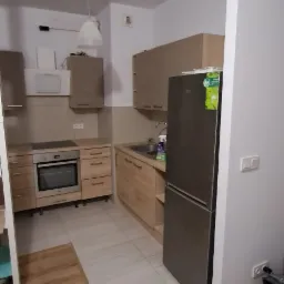 Jasna kuchnia z drewnianymi szafkami, sprzętem AGD i dwoma rodzajami podłóg: panelami imitującymi drewno oraz jasnymi płytkami.