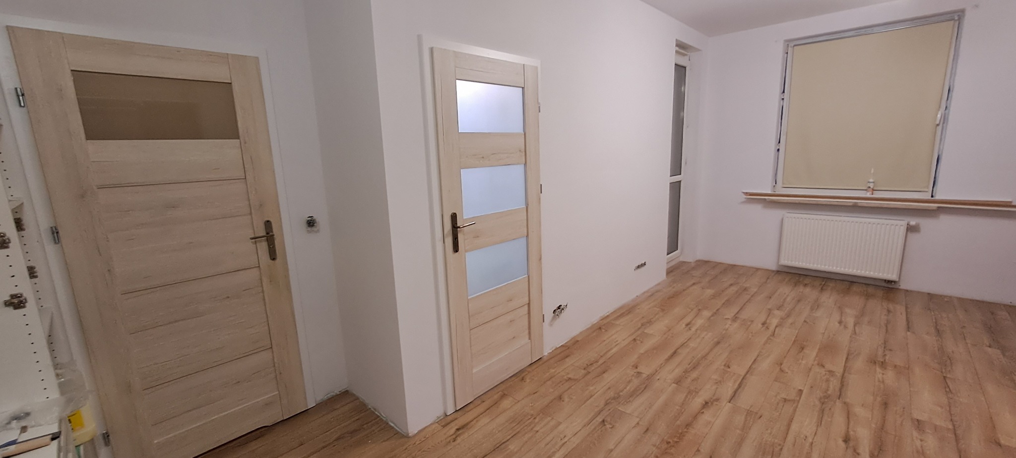 Wnętrze pokoju po remoncie z dwiema zamontowanymi, drewnopodobnymi drzwiami z przeszkleniami, panelami podłogowymi i grzejnikiem pod oknem z roletą.