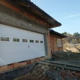 Biała brama garażowa segmentowa z małymi okienkami, zamontowana w surowym stanie budynku z cegły, widoczne materiały budowlane i przygotowania do wykończenia.