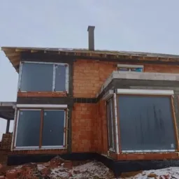 Dom w trakcie budowy z zamontowanymi oknami i roletami zewnętrznymi, widoczne cegły i elementy konstrukcyjne.