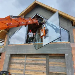Montaż okna o nietypowym, tr&oacute;jkątnym kształcie przy użyciu dźwigu przyssawkowego; widoczni dwaj monterzy w trakcie pracy na tle surowego stanu budynku.