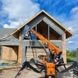 Montaż okna z użyciem specjalistycznego dźwigu przyssawkowego na placu budowy domu jednorodzinnego w stanie surowym otwartym, widoczna drabina oparta o ścianę.