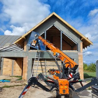 Montaż okna z użyciem specjalistycznego dźwigu przyssawkowego na placu budowy domu jednorodzinnego w stanie surowym otwartym, widoczna drabina oparta o ścianę.
