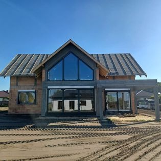 Dom w trakcie budowy z dużymi przeszkleniami i szarymi ramami, widoczne ślady opon na ziemi, błękitne niebo.