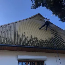 Pracownik w uprzęży myje metalowy dach myjką ciśnieniową, widoczne różnice w czystości powierzchni, zarys budynku poniżej.