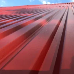 Czerwony, blaszany dach z widocznymi pofalowaniami, sfotografowany pod kątem, z fragmentem błękitnego nieba w tle.