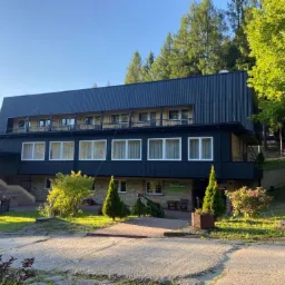 Dwupiętrowy budynek z ciemnym, pionowo żebrowanym dachem i jasnymi oknami, otoczony zielenią drzew i krzewów, widziany z poziomu ulicy w słoneczny dzień.