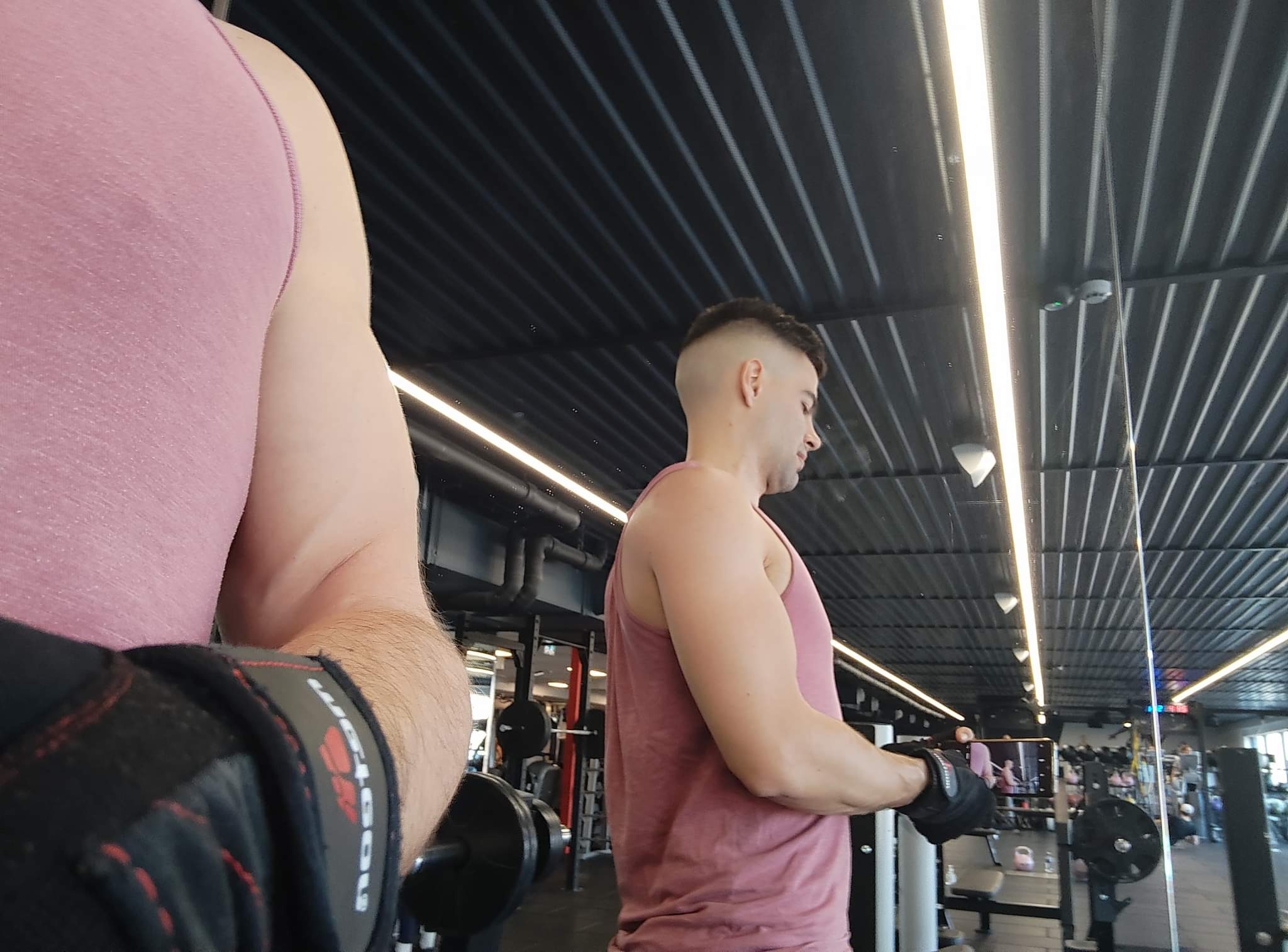 Mężczyzna w różowym tank topie ćwiczy biceps na siłowni w Wadowicach, widoczne obciążenia i lustro. Ujęcie z bliska na rękę z rękawicą.