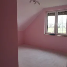 Pokój na poddaszu po malowaniu na różowo, widoczny fragment ściany w innym odcieniu różu, surowe wykończenie podłogi, okno z roletami.