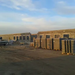 Wznoszenie ścian budynku z bloczków betonowych, palety z bloczkami na pierwszym planie, samochód dostawczy w tle, plac budowy.