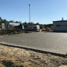 Świeżo ułożona, szara kostka brukowa na dużej powierzchni z widocznymi obrzeżami, otoczona piaskiem i gruzem, w tle budynki gospodarcze, wieża telekomunikacyjna i przyczepa kempingowa.