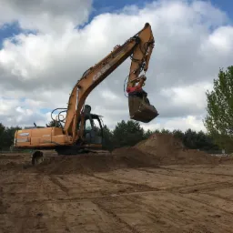 Żółta koparka CASE CX210 w Młodzieszynie podnosi łyżką ziemię na tle błękitnego nieba z chmurami, przygotowując teren pod budowę.