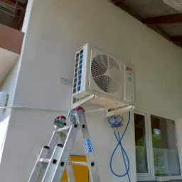 Biały klimatyzator zewnętrzny zamontowany na ścianie budynku, podłączony do manometrów za pomocą niebieskich przewodów, widoczna aluminiowa drabina Krause z naklejką ostrzegawczą 'GAZ'.