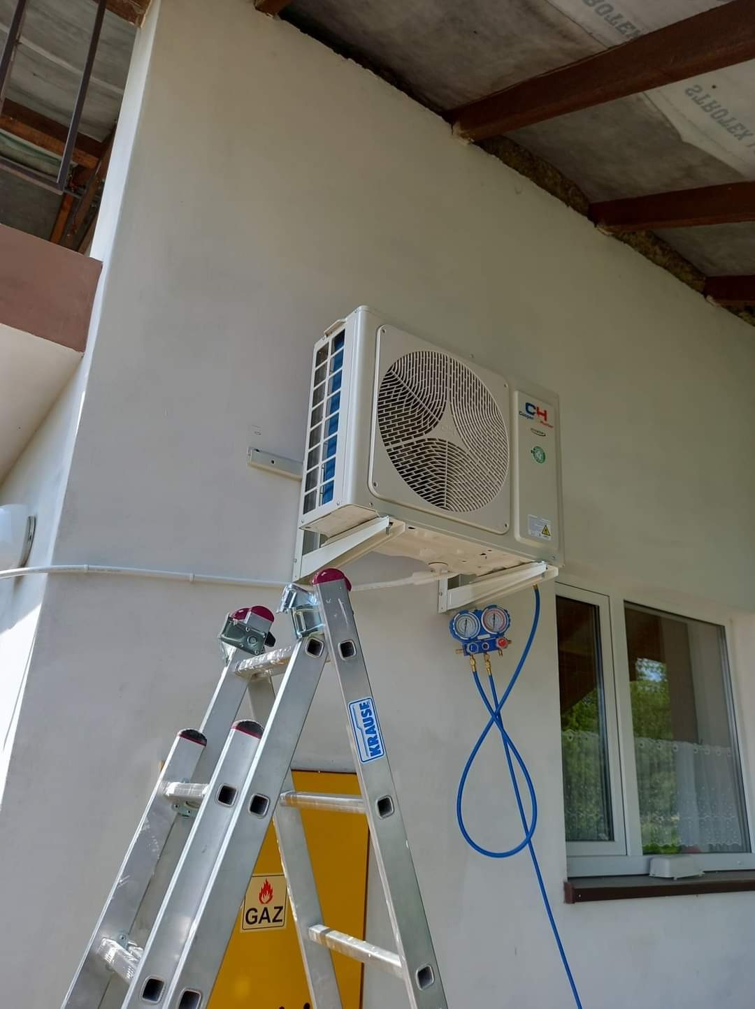 Biały klimatyzator zewnętrzny zamontowany na ścianie budynku, podłączony do manometrów za pomocą niebieskich przewodów, widoczna aluminiowa drabina Krause z naklejką ostrzegawczą 'GAZ'.