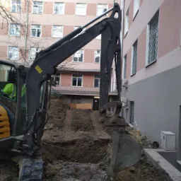 Żółto-czarna koparka na gąsienicach wykonuje wykop w ziemi, widoczny budynek z oknami z kratami w tle.