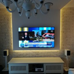 anteny serwis - Telewizor LCD zawieszony na ścianie z cegły, podświetlony taśmą LED, z widocznymi głośnikami po bokach i meblem RTV poniżej.