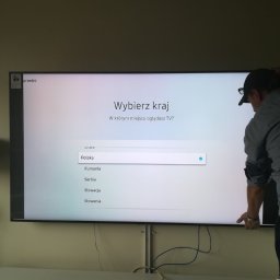 anteny serwis - Mężczyzna w czapce instaluje duży telewizor, na ekranie menu wyboru kraju, widoczne kable i fragment lampy.